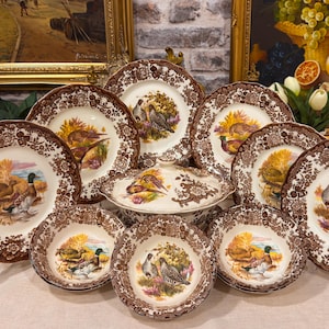 Op de afbeelding: Een set crèmekleurig keramisch servies met bruine bloemenranden en centrale illustraties van diverse wilde vogels, waaronder eenden en fazanten, bestaande uit meerdere borden, kommen en een serveerschaal met deksel.