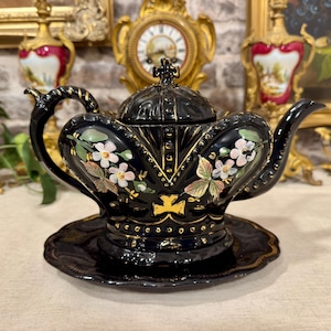 May include: A black ceramic teapot with a matching saucer, featuring a crown-shaped lid and body. The teapot is decorated with hand-painted white and pink flowers, green leaves, and gold accents along the edges and base, including a small gold cross.