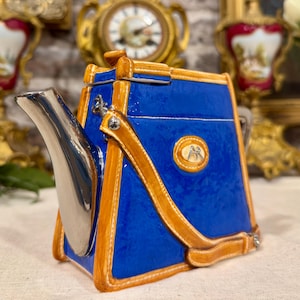 Può includere: Una teiera in ceramica a forma di borsa blu con finiture color cuoio e un beccuccio argentato. La teiera presenta un corpo rettangolare strutturato, un manico superiore e un piccolo emblema ovale sul lato con le lettere AR in oro.