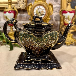 Può includere: Una teiera in ceramica nera con base quadrata abbinata, caratterizzata da finiture dorate e intricati motivi floreali verdi e oro. La teiera presenta un manico curvo, un beccuccio appuntito e un coperchio con una piccola impugnatura dorata.