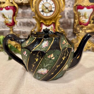 Peut inclure: Une théière en céramique avec un corps cannelé en spirale alternant des panneaux noirs et vert foncé. Les sections vertes sont ornées de motifs floraux peints à la main en rose, blanc et or, tandis que l'anse et le bec sont noirs.