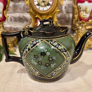 May include: A ceramic teapot featuring a matte olive green body with a black handle, spout, and lid. The center displays a diamond-shaped panel decorated with small black, white, and blue floral patterns outlined in gold, with black fluted details.