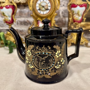 Puede incluir: Una tetera de cerámica negra con un medallón ovalado central que muestra la ilustración de un ciervo dorado, rodeado por un borde de puntos azul claro. El cuerpo está decorado con motivos florales y follaje dorados, con tapa y asa a juego.