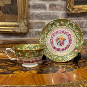 Op de afbeelding: Een porseleinen theekop met bijpassende schotel, voorzien van een saliegroene rand met gouden filigraan, een centraal bloemenboeket en een ring van roze rozen. De kop heeft een witte voet en een goudkleurig oortje, met kleine fruitmotieven op de schotel.