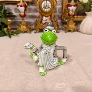 Peut inclure: Une théière en céramique en forme de grenouille verte portant un haut-de-forme gris, une veste grise et un pantalon blanc à rayures grises. La grenouille est assise avec une jambe étendue et le bec verseur ressemble à une pile de tasses.
