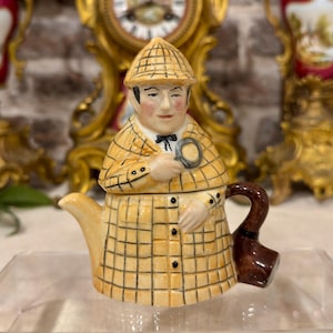 May include: A ceramic teapot shaped like a detective wearing a yellow and black plaid coat and a matching deerstalker hat. The figure holds a magnifying glass in one hand, features a black bow tie, and has a dark brown handle shaped like a pipe.