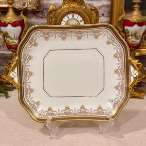 Peut inclure: Un plateau de service carré en porcelaine aux coins arrondis avec des poignées dorées. Le centre est blanc avec une fine bordure dorée. Le bord présente un motif décoratif de fleurs roses, jaunes et vertes sur un fond crème.