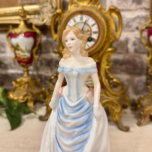 以下が含まれることがあります： ライトブラウンの髪をした人物の磁器製フィギュアで、肩を出したガウンを着用しています。ドレスは主に白で、水色の縦のアクセントとウエストにピンクのディテールが施されています。フィギュア全体が滑らかで光沢のある仕上がりです。