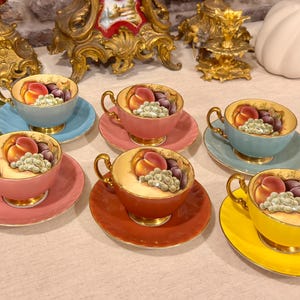 Pode incluir: Um conjunto de seis chávenas de chá e pires em porcelana em cores variadas, incluindo azul claro, rosa, azul-petróleo, laranja e amarelo. Cada chávena apresenta um design pintado à mão de um pêssego, uvas e ameixas com bordas douradas.