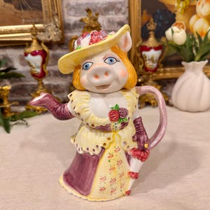 Könnte beinhalten: Eine Keramik-Teekanne in Form einer Schweinefigur mit einem gelben Blumenhut, einem gerüschten gelben Oberteil mit Rosenakzenten und einem violetten Rock. Die Figur hat orangefarbenes Haar, blaue Augen und hält einen kleinen rosa Regenschirm.
