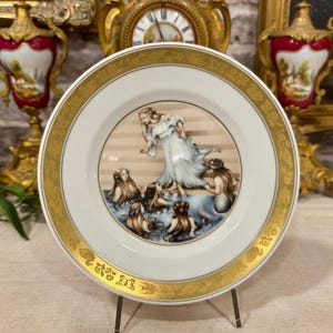 Può includere: Un piatto decorativo in porcellana bianca con un'illustrazione centrale di una donna in un abito bianco fluente vicino a un gruppo di sirene in acqua blu. Il bordo largo è dorato e decorato con motivi floreali e di cigni.