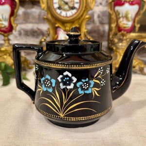 May include: A glossy black ceramic teapot featuring a hand-painted floral design with light blue and white petals, accented by gold stems and leaves. The teapot includes a matching lid, a curved handle, and a spout, all detailed with gold trim.