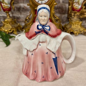 Op de afbeelding: Een keramische theepot in de vorm van een ouder persoon in een roze jurk met kleine blauwe bloemaccenten, een witte kraag met ruches, een roze cape en een blauwe muts. De figuur houdt een blauwe paraplu vast en heeft een gebogen handvat.