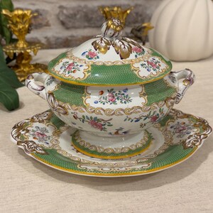 May include: A porcelain tureen with a matching saucer, featuring a white base with green textured borders, gold trim, and colorful floral patterns. The lidded bowl has two side handles and a decorative finial on top, showcasing intricate painted details.