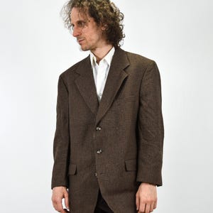 May include: A brown blazer with a textured weave, featuring a notched lapel and three buttons. The blazer has two front pockets and is worn over a white collared shirt. The jacket is a classic style.