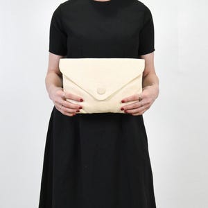 Könnte beinhalten: Eine cremefarbene Umschlag-Clutch mit Knopfverschluss. Die Clutch wird vor einer Person getragen, die ein schwarzes Kleid trägt. Der Hintergrund ist schlicht weiß.