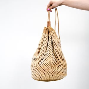 Op de afbeelding: Een beige trekkoordtas met een geweven netontwerp. De tas heeft een houten tuimelsluiting en een trekkoord van touw. De tas wordt door een hand tegen een witte achtergrond omhoog gehouden. De tas is lichtbruin.
