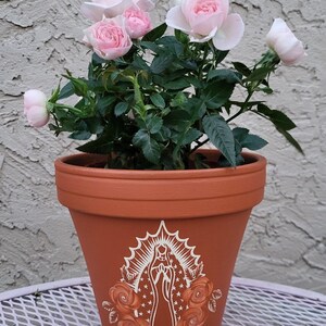 May include: A terracotta flower pot featuring a white illustration of a religious figure and roses. The pot holds a rose plant with pink flowers and green foliage. The pot is on a white metal table.