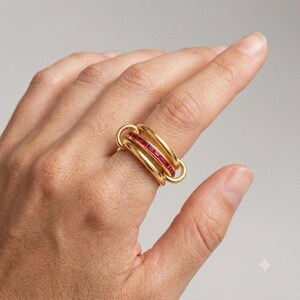 May include: A gold ring featuring three interconnected bands. The central band is set with a row of small, square-cut red gemstones, while the two outer bands are smooth, polished gold. The design creates a layered, multi-band appearance.