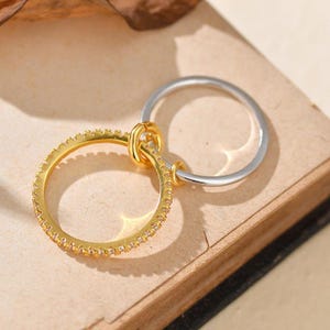 Può includere: Un anello color oro con una fascia di piccole pietre trasparenti è collegato a un anello color argento. Gli anelli sono collegati da un piccolo connettore color oro. Gli anelli sono esposti su una superficie marrone.