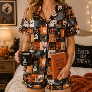 May include: A short-sleeve button-down pajama shirt and matching shorts set featuring a patchwork pattern of orange, black, and white Halloween motifs including ghosts, pumpkins, bats, and floral designs. A black mug with the text HEY BOO is held.