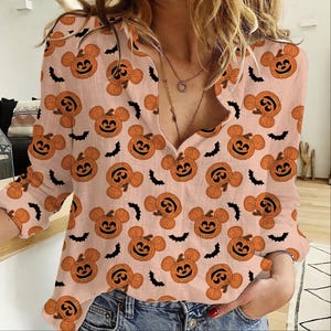 May include: A light peach-colored long-sleeve button-down shirt featuring an all-over print of orange jack-o'-lanterns with mouse-shaped ears and small black bats. The shirt has a V-neckline and is worn with blue denim jeans.