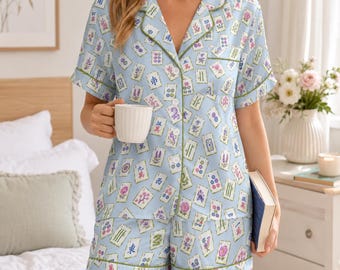 May include: A light blue short-sleeve pajama set featuring a repeating pattern of mahjong tiles and floral illustrations. The button-down top has a notched collar with green piping, and the matching shorts also feature green piping along the hem.