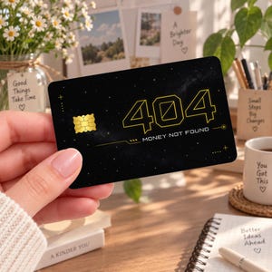 May include: A black rectangular credit card featuring a starry night sky design with a gold chip on the left. The card displays the yellow outlined text 404 and the phrase MONEY NOT FOUND in a modern font, accented with thin yellow lines and plus signs.
