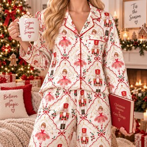 May include: A white button-down pajama set featuring a repeating pattern of nutcrackers and ballerinas in red, green, and gold. The pajamas have red piping along the collar and front. A white mug with the text Sugar Plum Season and a red book are held.