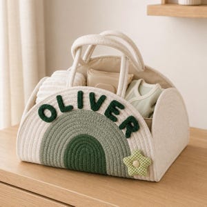 May include: A cream-coloured rope storage basket with two handles, featuring a green rainbow design on the front. The name OLIVER is stitched in dark green letters across the rainbow. A small green and white checkered star is attached to the side.