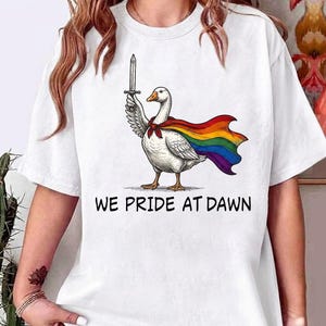 Pode incluir: Uma t-shirt branca de manga curta com um gráfico de um ganso branco que usa uma capa com as cores do arco-íris e segura uma espada. Abaixo do ganso, o texto WE PRIDE AT DAWN está escrito em letras maiúsculas pretas no peito.
