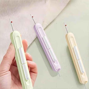 May include: Three seam rippers with retractable blades, each featuring a pastel-colored plastic handle in light green, lavender, or cream. Each tool has a white sliding mechanism on the front and a small red safety ball at the tip of the metal blade.