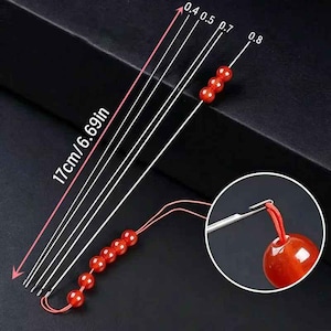 May include: A set of four silver-colored beading needles with hooked ends, each measuring 17 cm or 6.69 inches in length. The needles are labeled with sizes 0.4, 0.5, 0.7, and 0.8. Red strings with small red beads are attached to the needles.