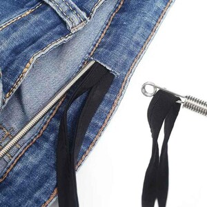 May include: A blue denim fabric waistband featuring a black drawstring threaded through a metal eyelet. A silver metal spring-loaded tool is shown pulling the black fabric drawstring through the denim channel for easy adjustment.
