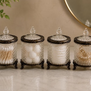May include: A set of four cylindrical glass jars with textured surfaces, each featuring a removable lid with a pointed finial and a dark bronze-toned metal base with ornate feet. The jars contain cotton swabs, cotton balls, and jewellery.