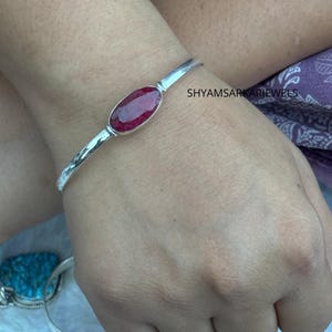 Puede incluir: Una pulsera de plata con una piedra preciosa de color rojo intenso facetada en forma ovalada en el centro. La banda de metal tiene un acabado pulido. El texto SHYAMSARKARJEWELS es visible en la esquina superior derecha de la imagen.