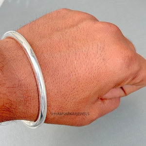 Puede incluir: Un brazalete de plata maciza pulida con una superficie lisa y redondeada. El metal tiene un acabado brillante y reflectante. El texto SHYAMSARKARJEWELS está impreso en pequeñas letras negras sobre la piel cerca del brazalete.