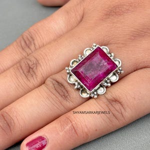 May include: A rectangular, deep pink gemstone ring set in a silver frame with decorative scalloped edges. The setting features small silver spheres around the perimeter of the stone. The text SHYAMSARKARJEWELS is visible on the silver band.
