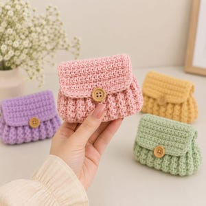 May include: A small, rectangular, crocheted pouch in light pink with a textured stitch pattern, a fold-over flap, and a single round wooden button closure. Similar pouches in purple, yellow, and light green are arranged in the background.