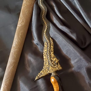 Puede incluir: Una daga de hoja ondulada con metal oscuro y filigrana dorada en la base. El mango es de madera marrón pulida con forma curva. Se acompaña de una vaina larga de color marrón claro con grabados intrincados de dragones en su superficie.