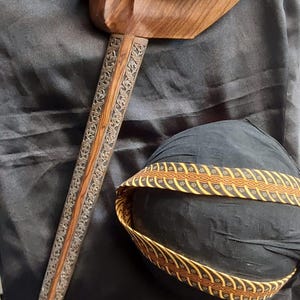 Op de afbeelding: Een traditionele Indonesische keris-dolk met een gebogen houten gevest, een donker metalen handvat en een houten schede met gedetailleerd zilverkleurig metaalfiligraan, vergezeld door een zwart stoffen hoofddeksel met een gele patroonband.