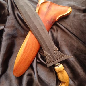 May include: A keris dagger with a dark, textured, wavy blade featuring intricate carvings at the base. The handle is crafted from light-colored wood with a curved, ergonomic shape. It is accompanied by a polished, reddish-brown wooden sheath.