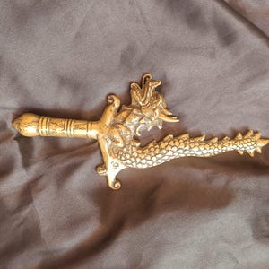 Puede incluir: Un abrecartas de metal dorado con forma de espada y motivo de dragón. El mango presenta un diseño cilíndrico con bandas horizontales, mientras que la hoja está formada por el cuerpo alargado y escamoso de un dragón con una cabeza detallada.