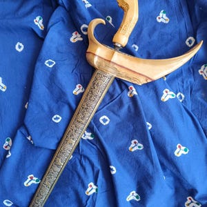 Puede incluir: Una daga keris con una empuñadura de madera clara con tallas intrincadas y una guarda transversal ancha y curva de madera. La vaina larga y recta es de metal oscuro y está decorada con detallados patrones en relieve de animales y follaje.