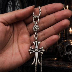 Pode incluir: Um colar de prata esterlina com um pendente em forma de cruz de braços alargados e um conector floral circular. A corrente é de estilo elo quadrado e o anel conector tem gravado o texto 925, indicando a pureza da prata.