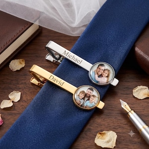 May include: Two metal tie clips, one silver-toned and one gold-toned, each featuring a circular photo insert and an engraved name. The silver clip is engraved with the name Michael, and the gold clip is engraved with the name David.
