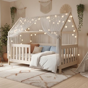 Può includere: Una struttura letto in legno chiaro a forma di casetta con doghe verticali, drappeggiata con un tessuto bianco semitrasparente e illuminata da una catena di luci a sfera calde. Il letto è coperto da un piumino bianco e cuscini blu e beige.