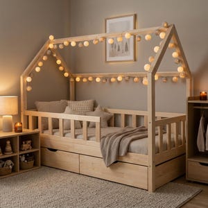 Peut inclure: Un cadre de lit en bois clair en forme de maison avec une barrière à lattes et deux tiroirs de rangement en dessous. La structure est décorée d'une guirlande lumineuse à boules. Le lit est garni d'oreillers et d'un plaid aux tons neutres.