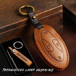 Puede incluir: Una funda de cuero color canela para llave de coche con costuras visibles, unida a una correa de cuero negro y una anilla de metal color latón. Una etiqueta de cuero canela con el texto grabado NAME 123456789 y un destornillador pequeño están en una caja negra.