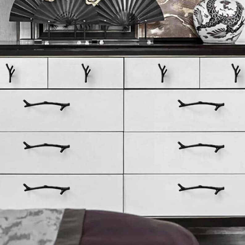 May include: A white dresser featuring eight drawers with matte black hardware. The top row has four small knob-style pulls shaped like twigs, while the three lower rows each have two long, horizontal branch-shaped handles on each drawer front.